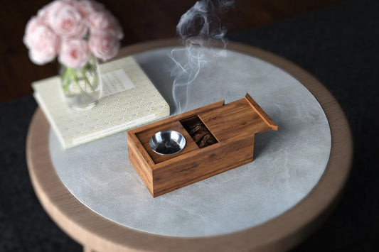 Wooden incense burner with smoke on a round stone surface, next to a vase of pink flowers and a book.